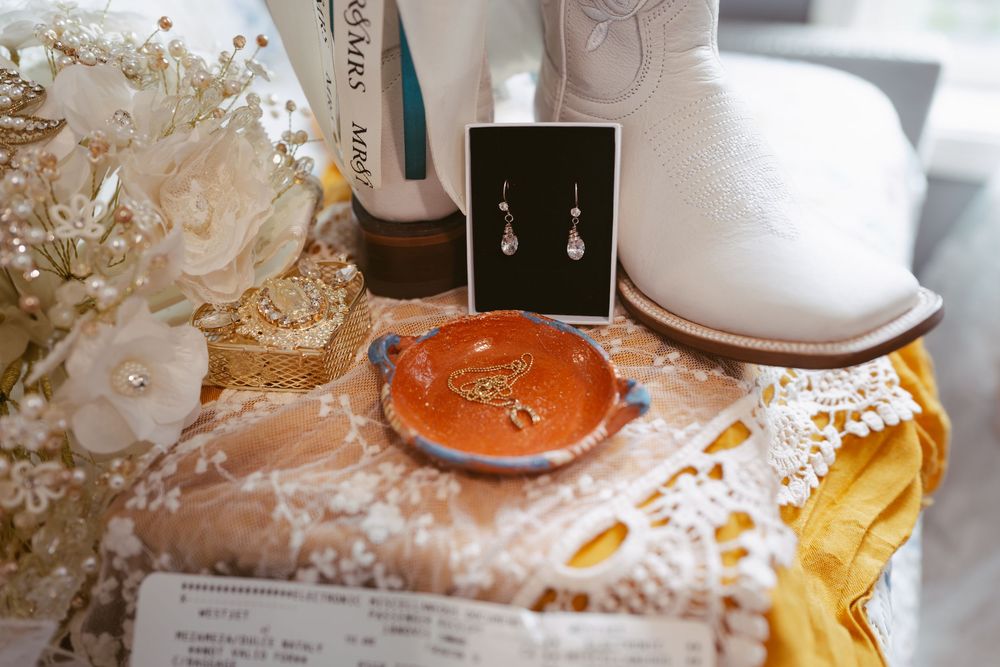 Wedding flat lay with white cowboy boots, drop earrings, orange ring dish, lace details, and bridal bouquet.