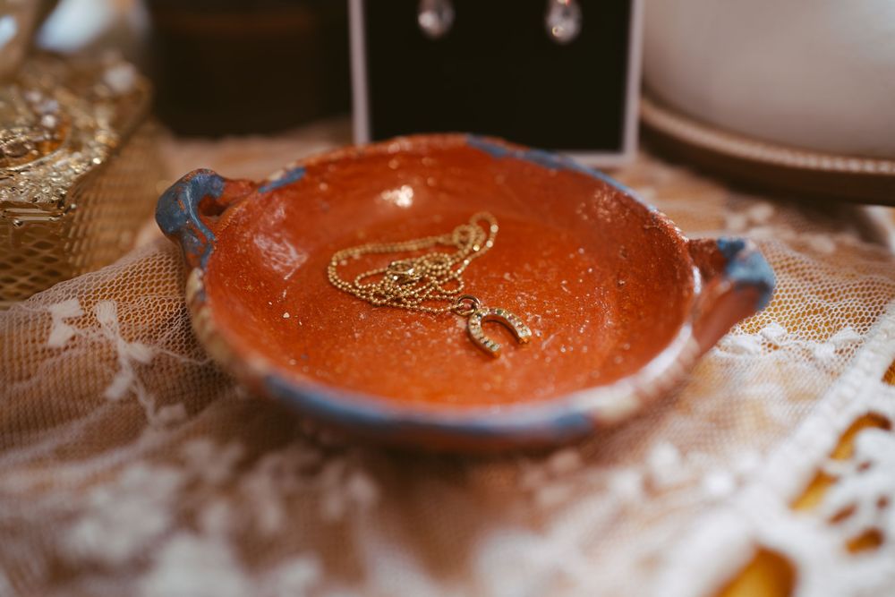 Orange ceramic trinket dish with gold bird jewelry piece, decorative glitter finish and handles on rustic lace surface.