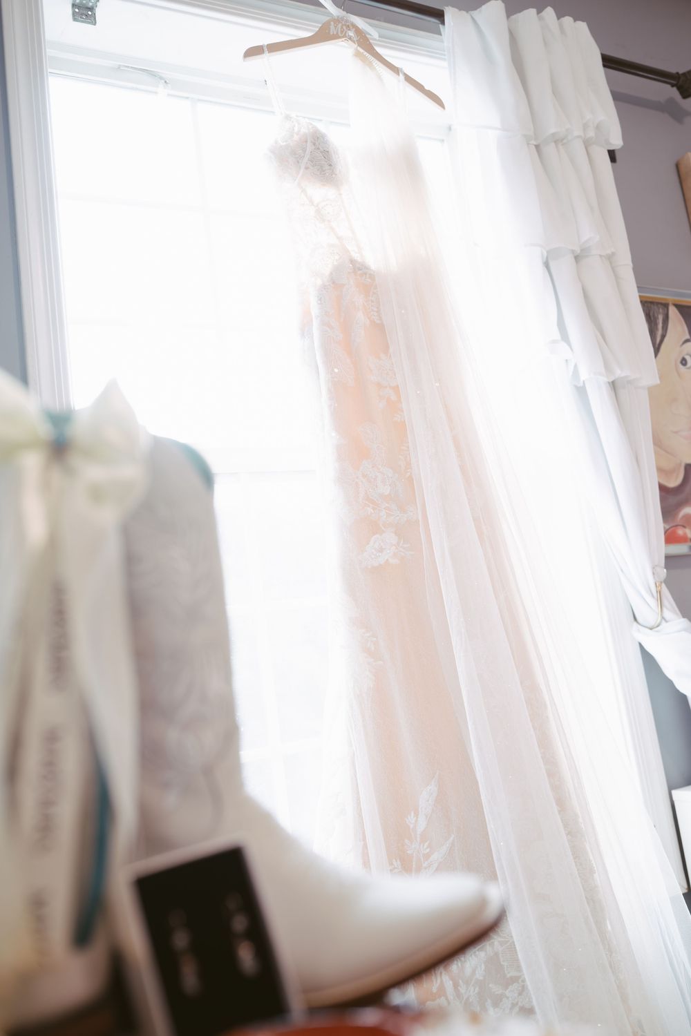 Elegant lace bridal gown hanging by window with white cowboy boots in foreground.