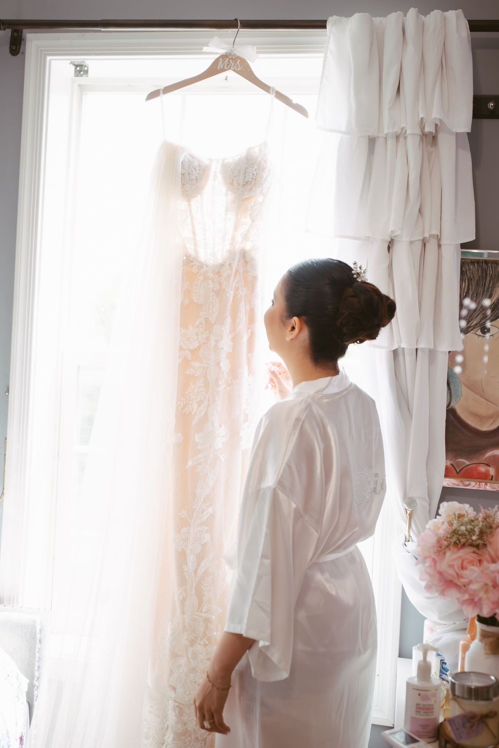 Bride in white robe admiring her lace wedding dress hanging in sunlit window.