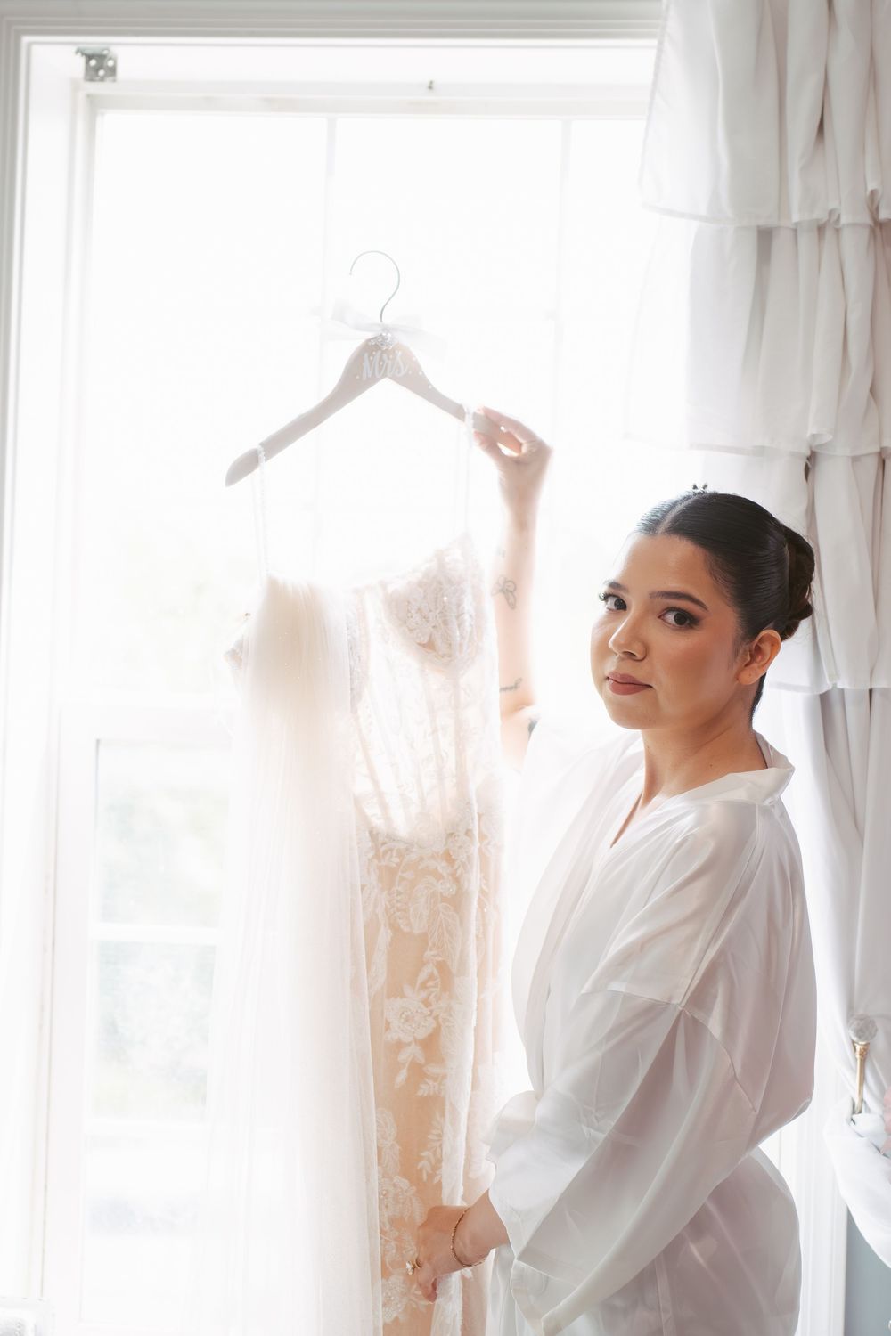 Bride in white robe holding lace wedding dress on hanger near bright window before ceremony.