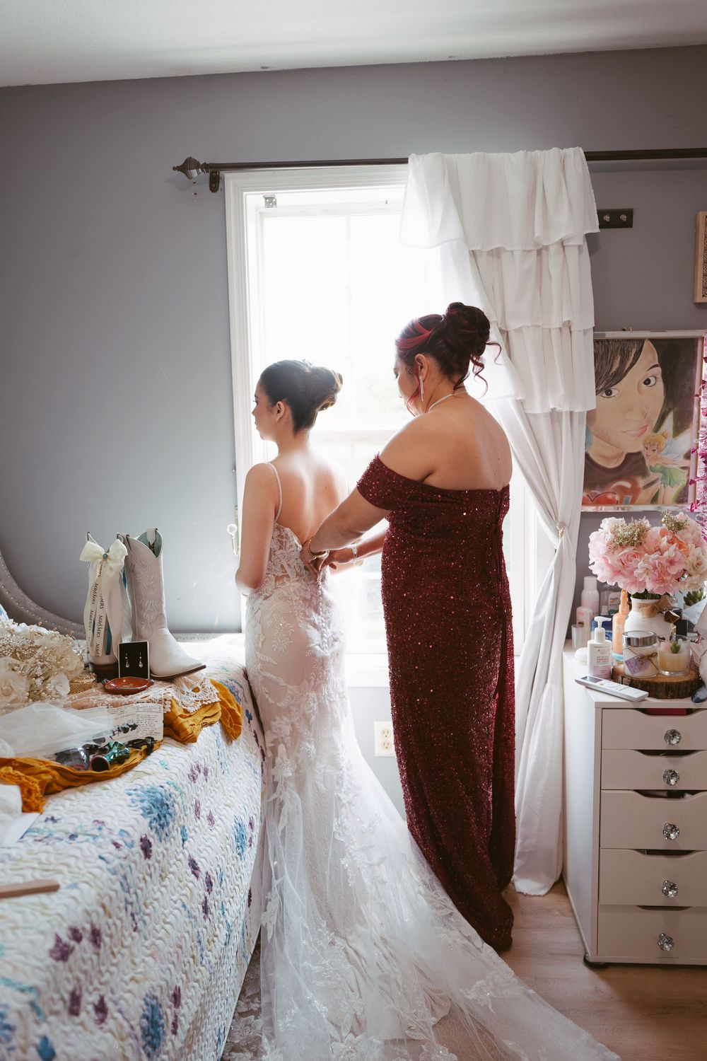 A helper fastens the back of a brides lace wedding gown in a softly lit bedroom before the ceremony.