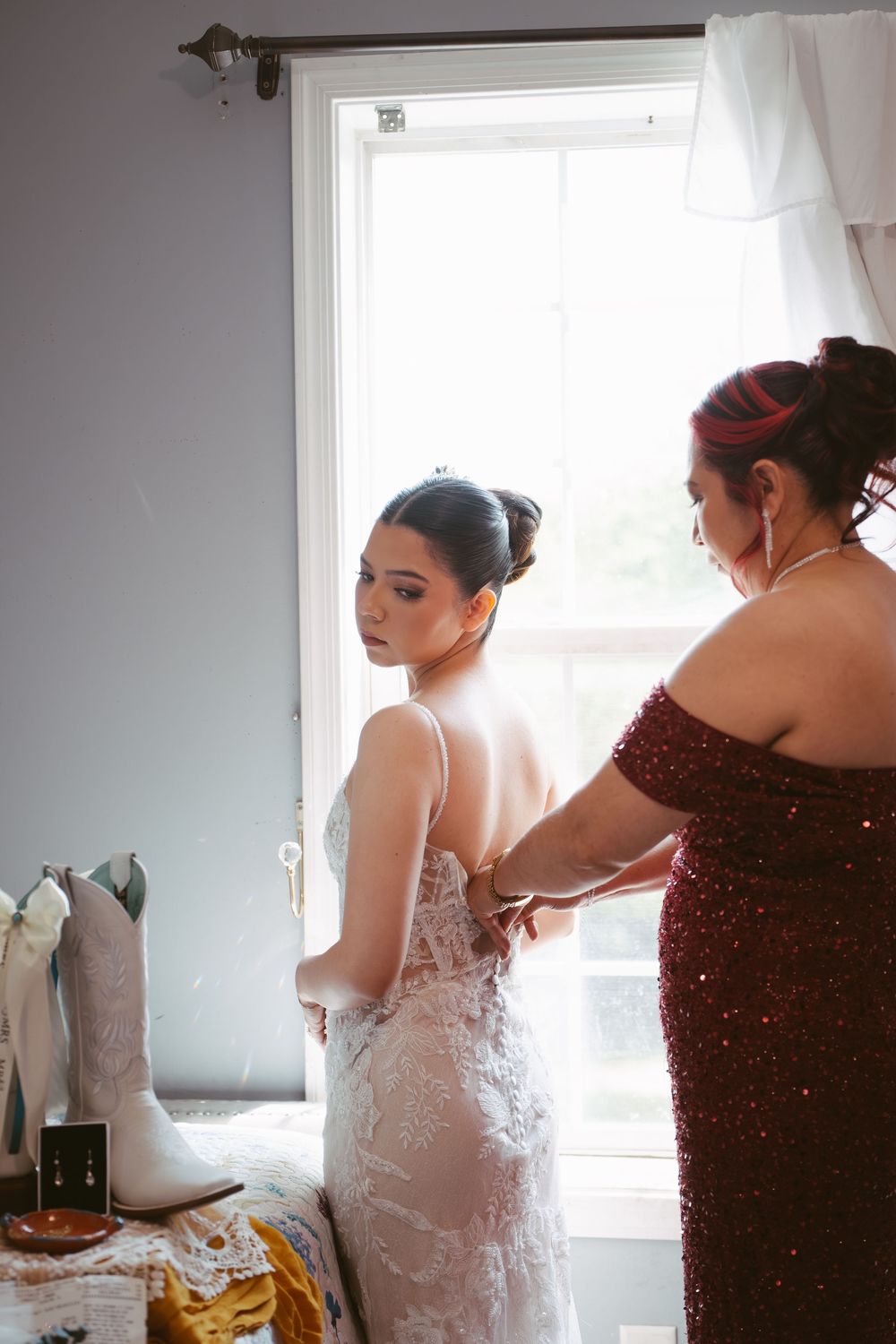Bride in lace wedding gown being helped by woman in burgundy dress near bright window with white cowboy boots nearby.
