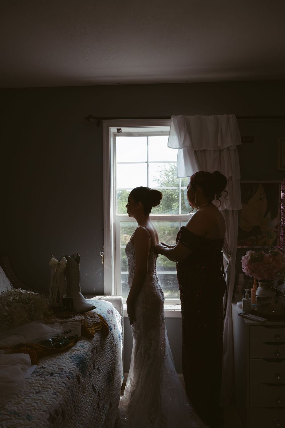 Silhouette of bride in wedding dress being helped by bridesmaid near bright window in dimly lit room.