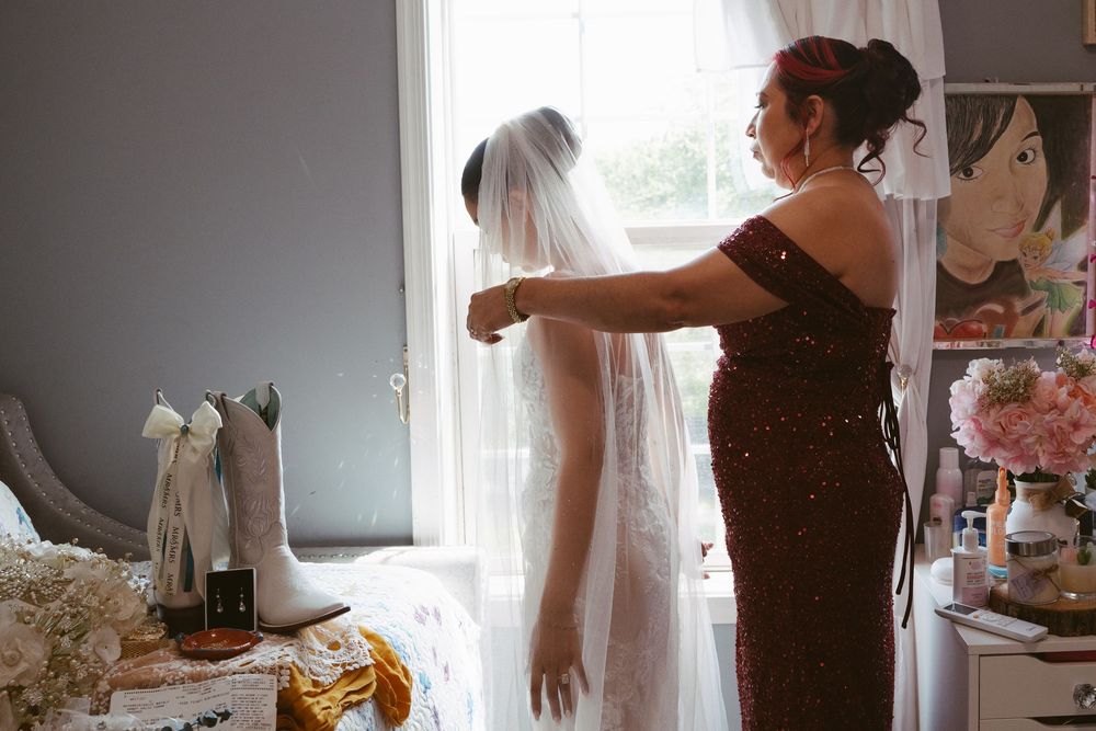 A mother helps her daughter adjust a bridal veil near a bright window, with white cowboy boots visible nearby.