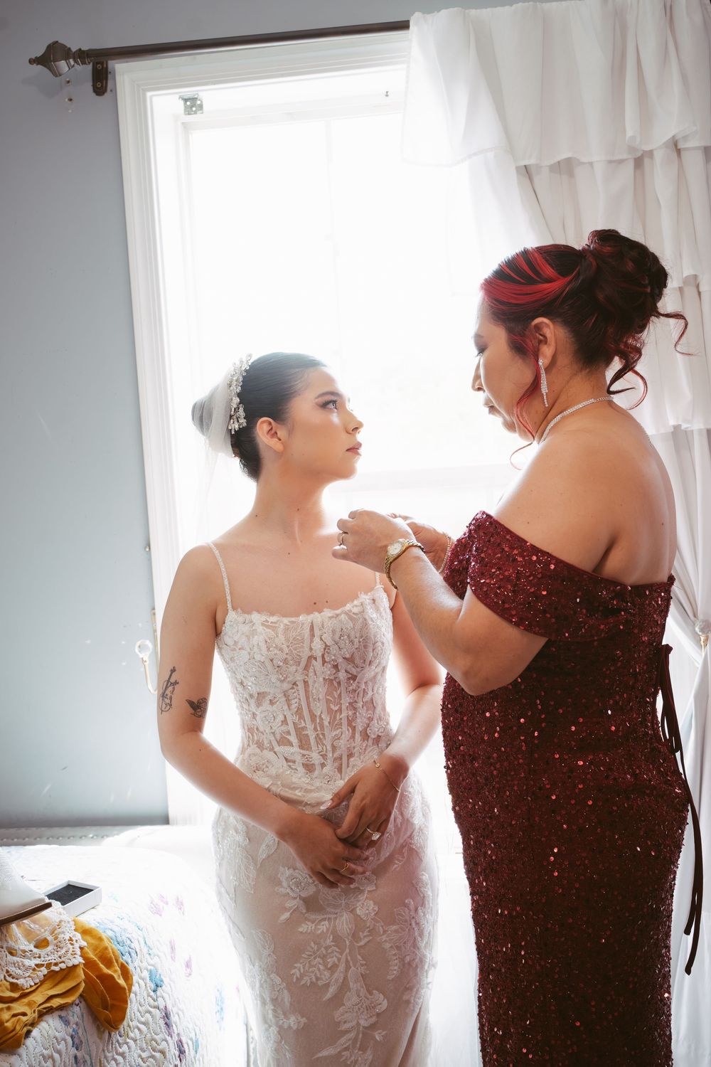 Bride in white lace gown gets help with necklace from woman in red sequin dress near bright window.