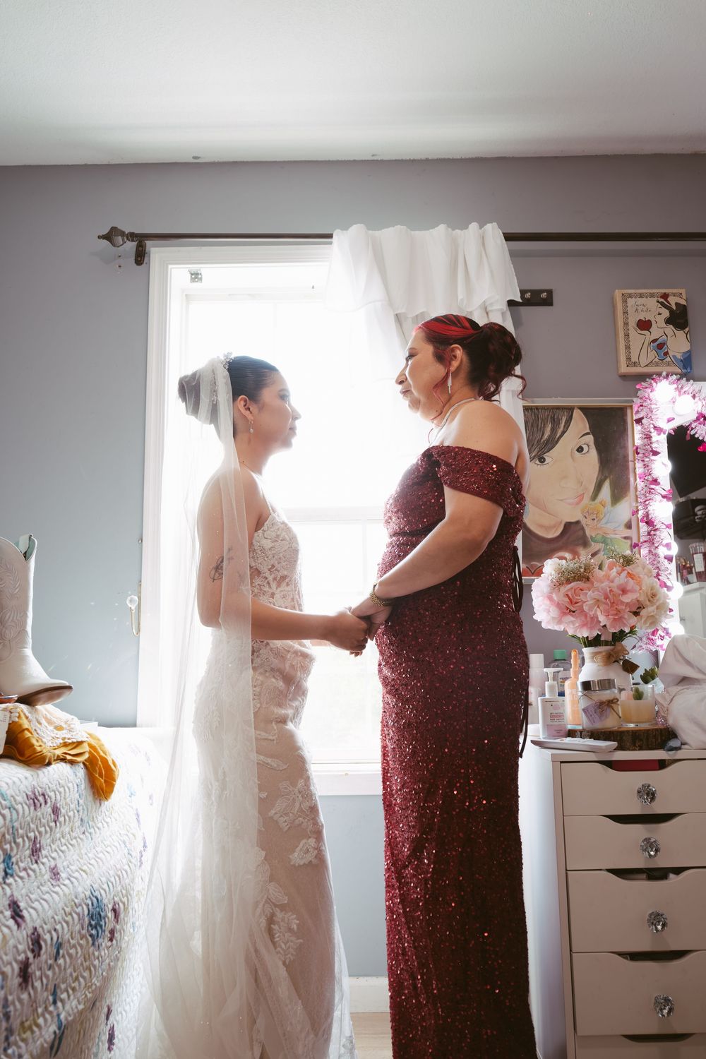 Bride in lace gown holding hands with mother in burgundy sequin dress by window.