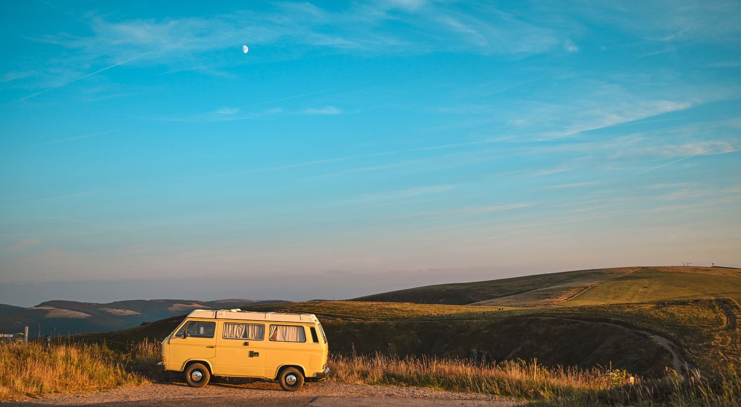 combi Volkswagen orange garé sur une route de crête, ciel bleu dégagé, collines dorées à perte de vue