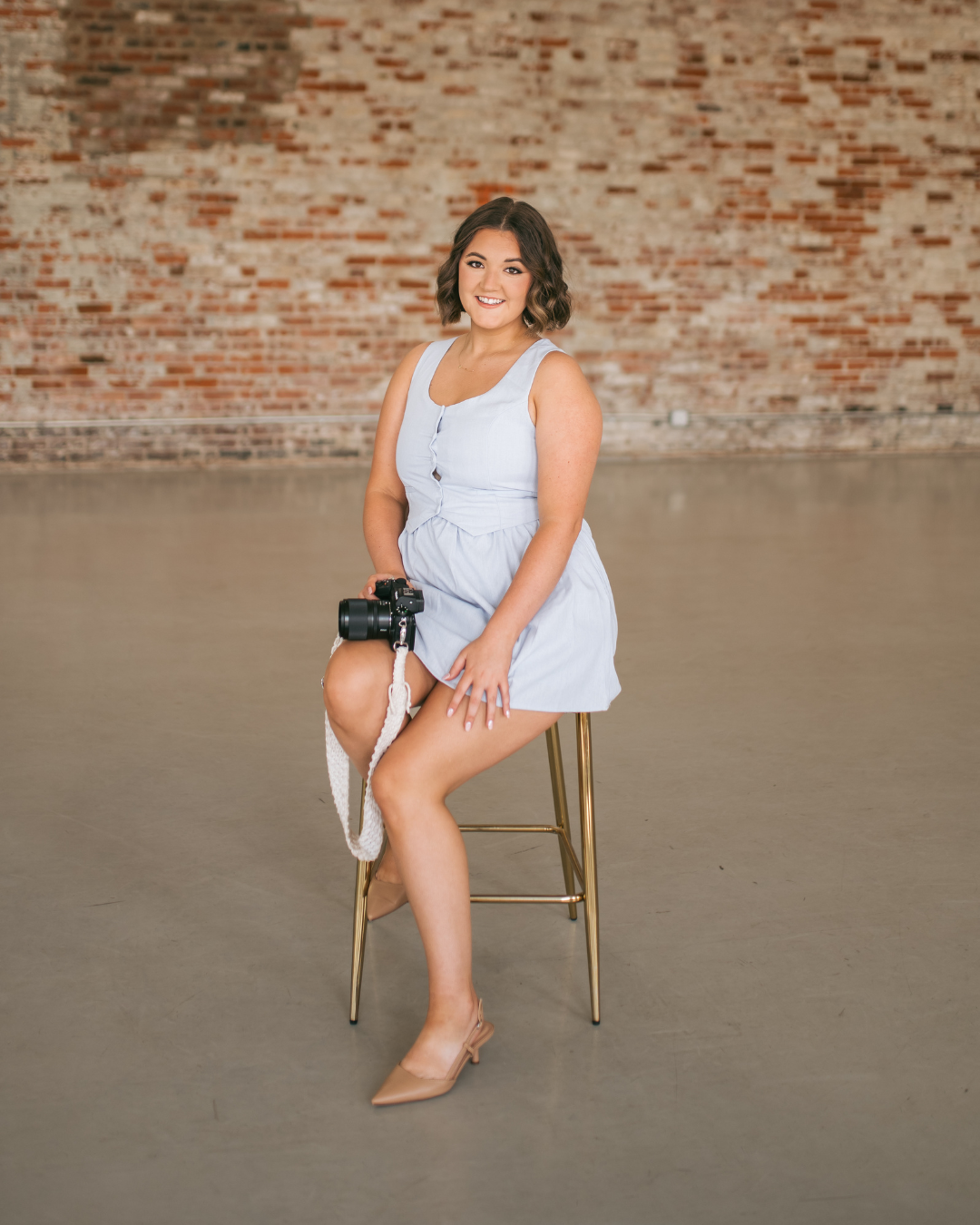 Young woman in light blue dress sits on gold stool holding camera in front of brick wall, luxury wedding photographer NC