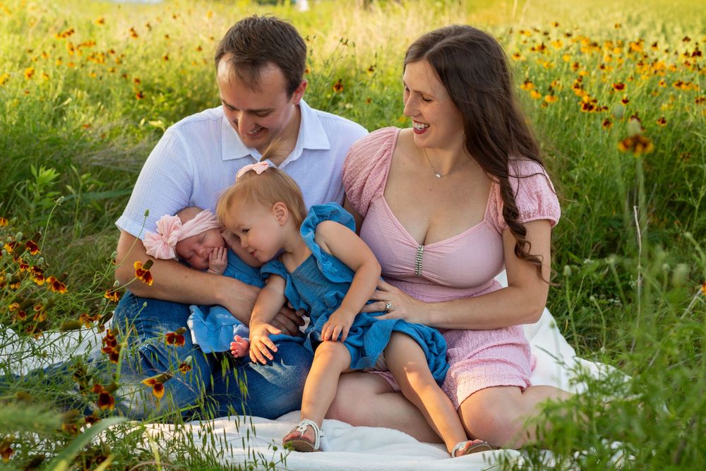 Family portrait of parents with two young children sitting on a blanket in a vibrant wildflower field.