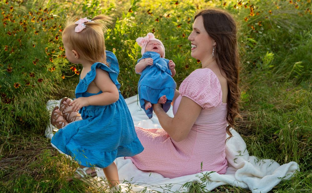 Mother in pink dress sits outdoors holding baby while toddler girl in blue dress plays in flower field.