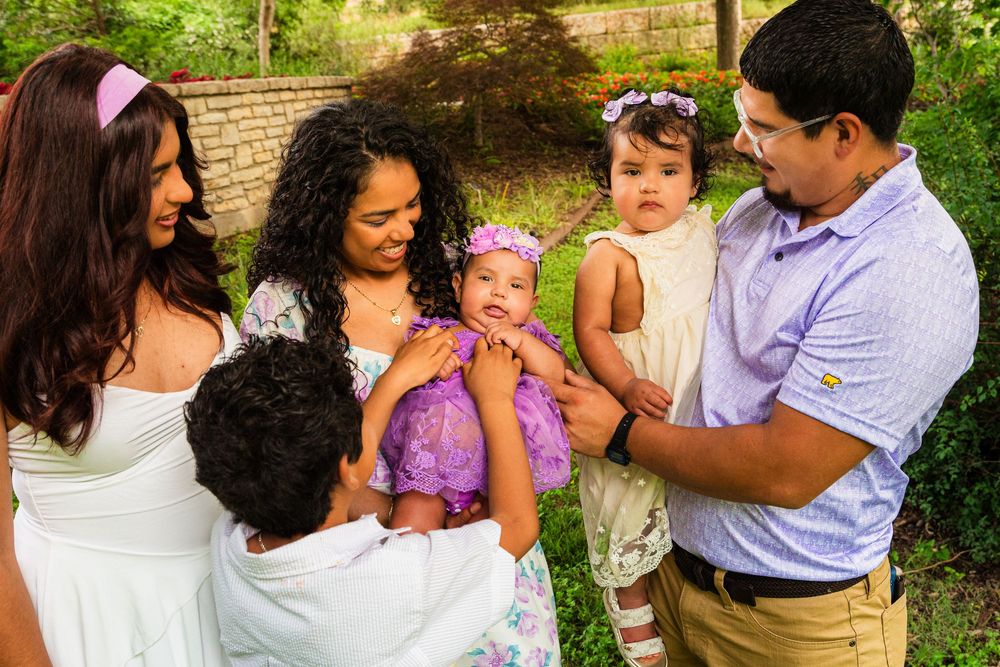 Family gathering outdoors, admiring a baby in purple outfit, surrounded by lush greenery.