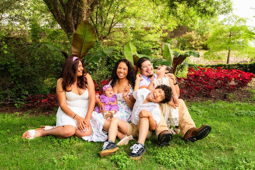 Happy family of five laughing and sitting together on green grass in a lush garden with red flowers.
