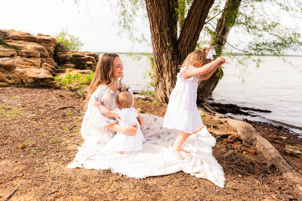 Mother in white dress sits with baby on blanket by lake as young girl in white dress plays near large tree.
