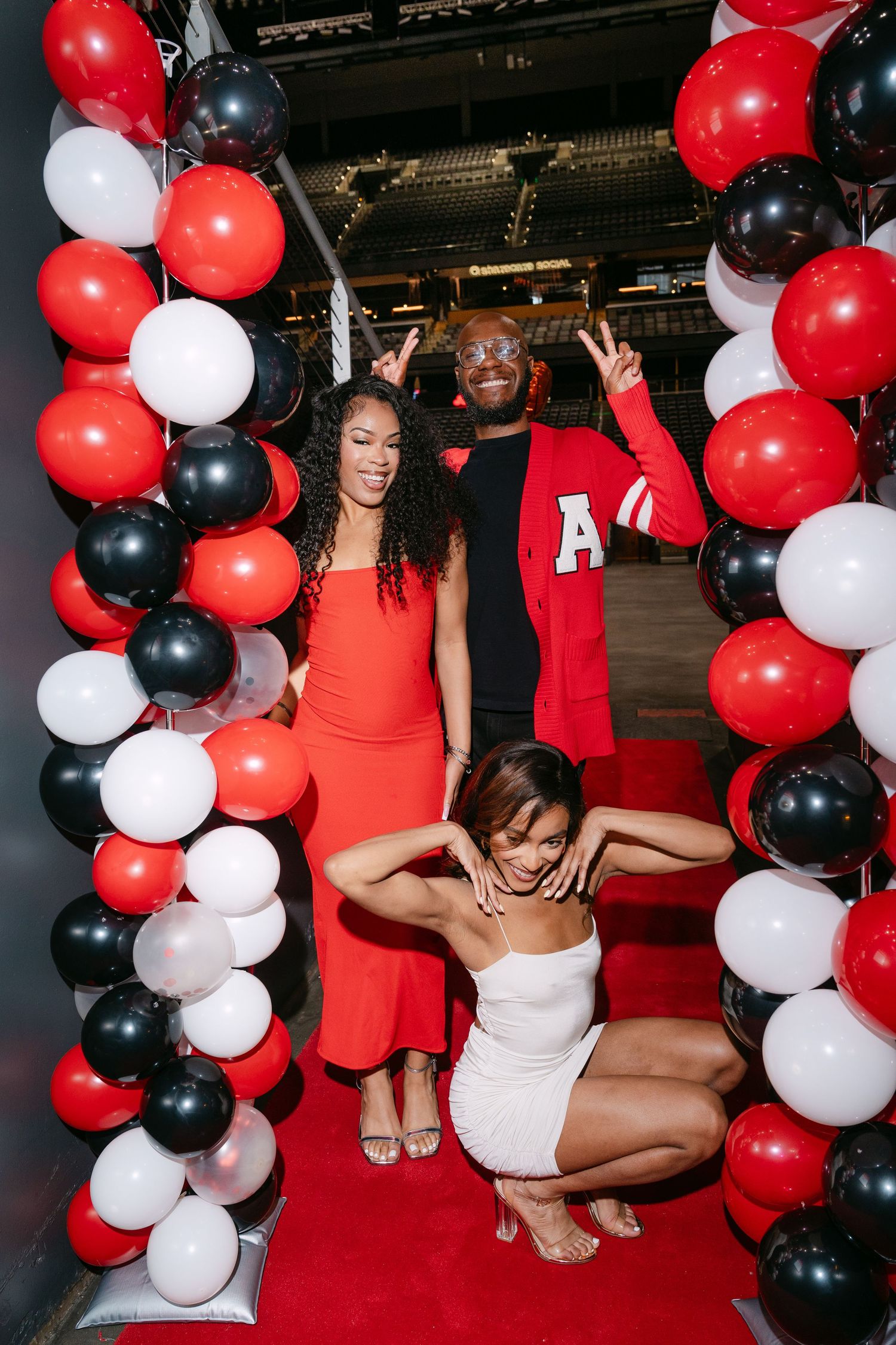 Three people posing in a red, black, and white balloon arch at a festive party celebration.