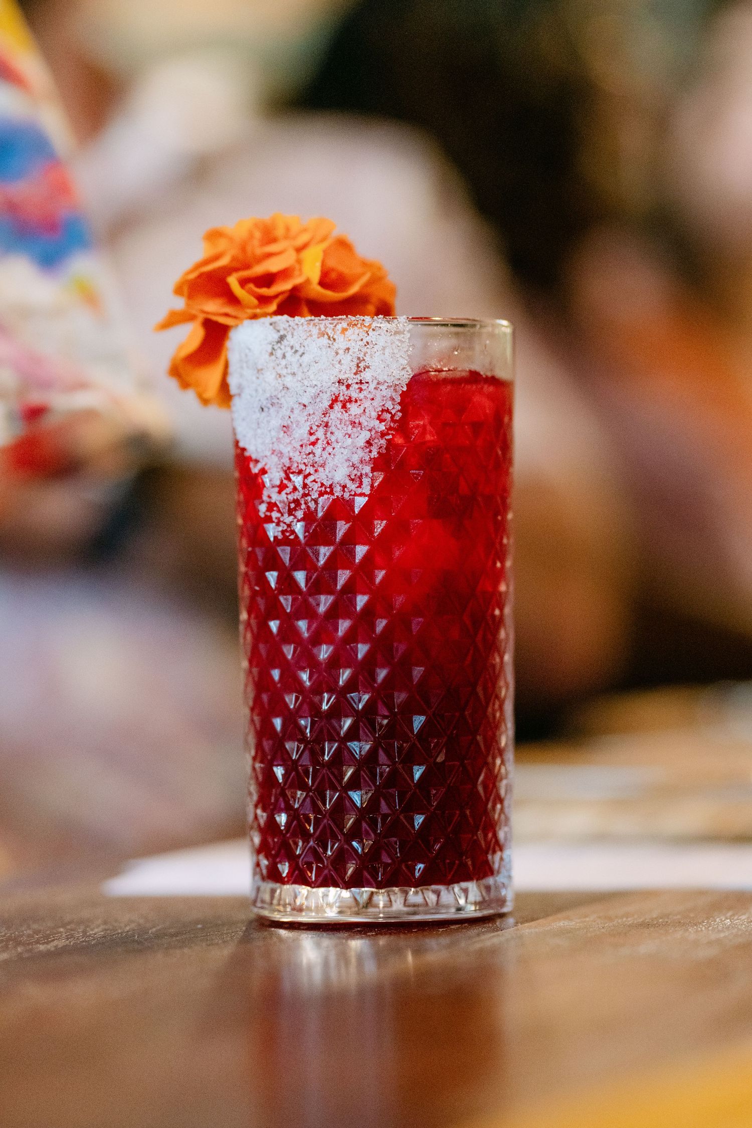 A vibrant red cocktail in a textured highball glass, topped with crushed ice and an orange flower garnish.