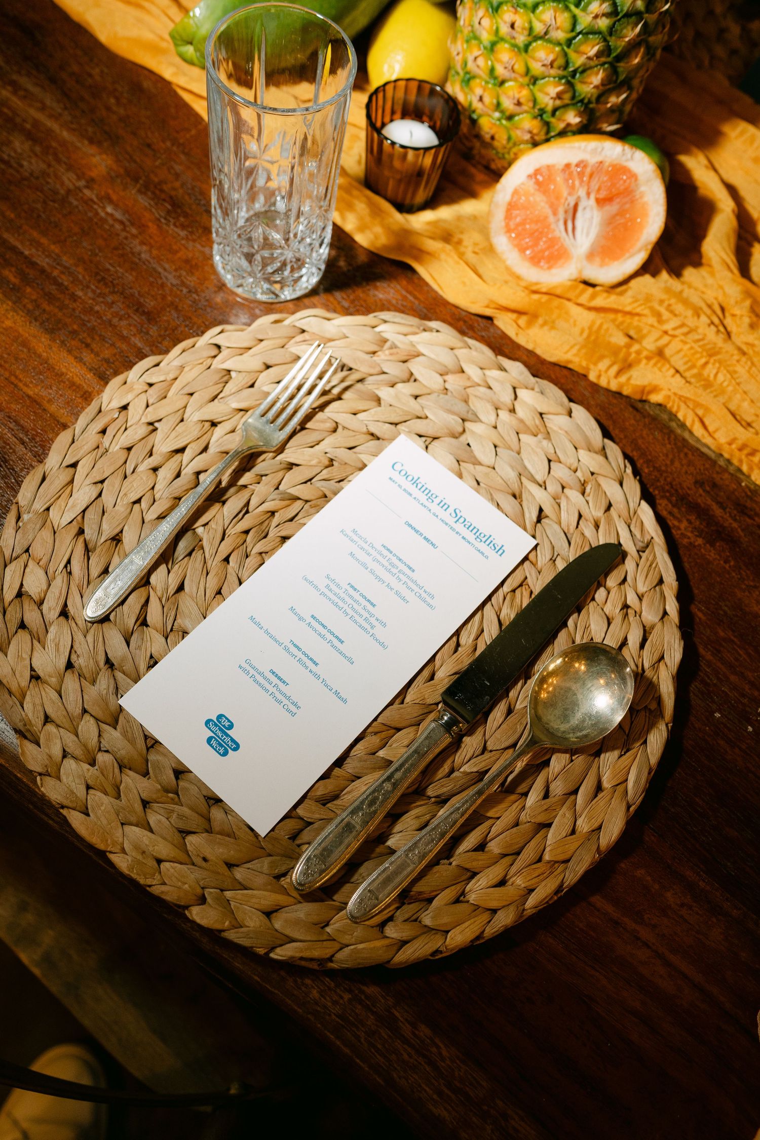 Woven placemat table setting with menu card, vintage cutlery, crystal glass, and tropical fruits on wooden table.