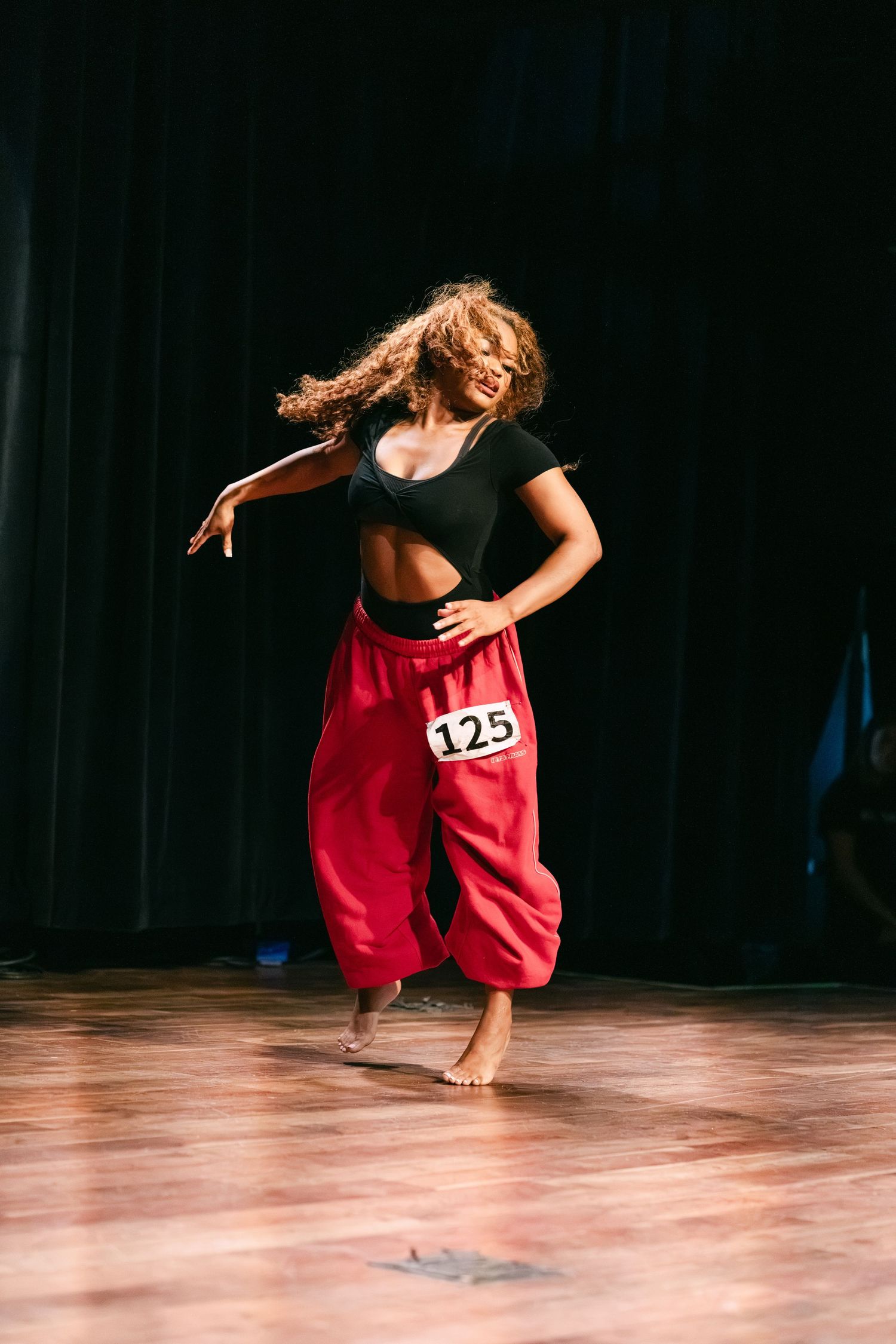 Dancer wearing red baggy pants and black crop top with number 125 performs barefoot on wooden stage.