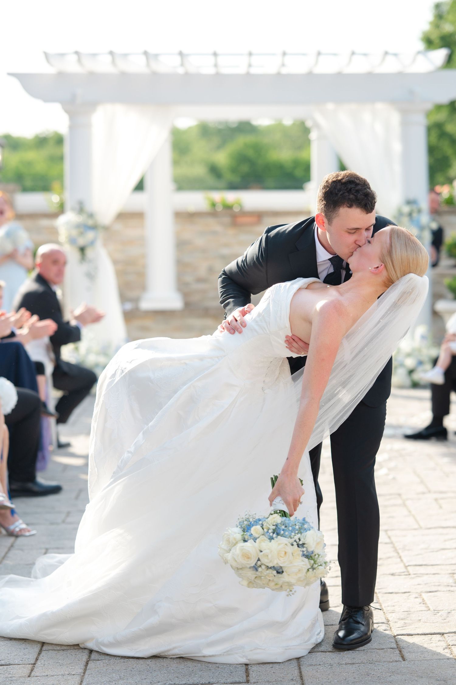A smiling bride and groom walk hand-in-hand along a garden path beside a white gazebo on their wedding day.