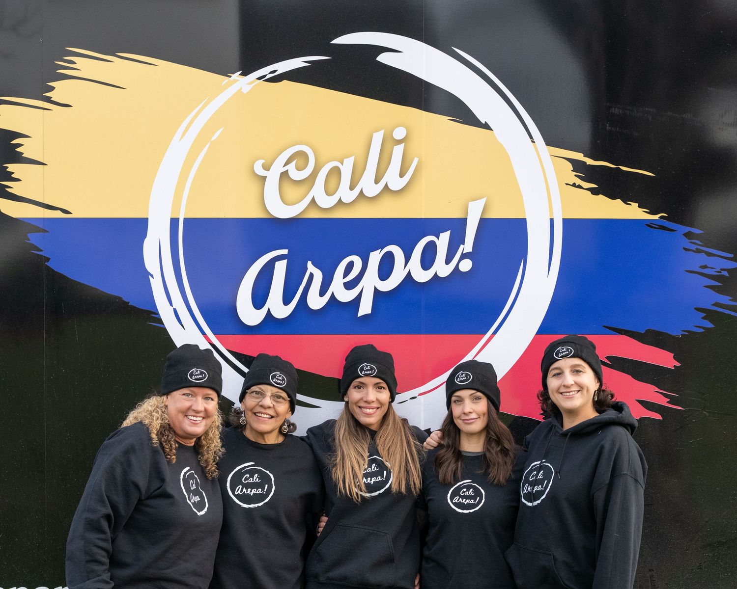 Five women in Cali Arepa branded black hoodies pose together in front of a Colombian flag-themed logo sign.