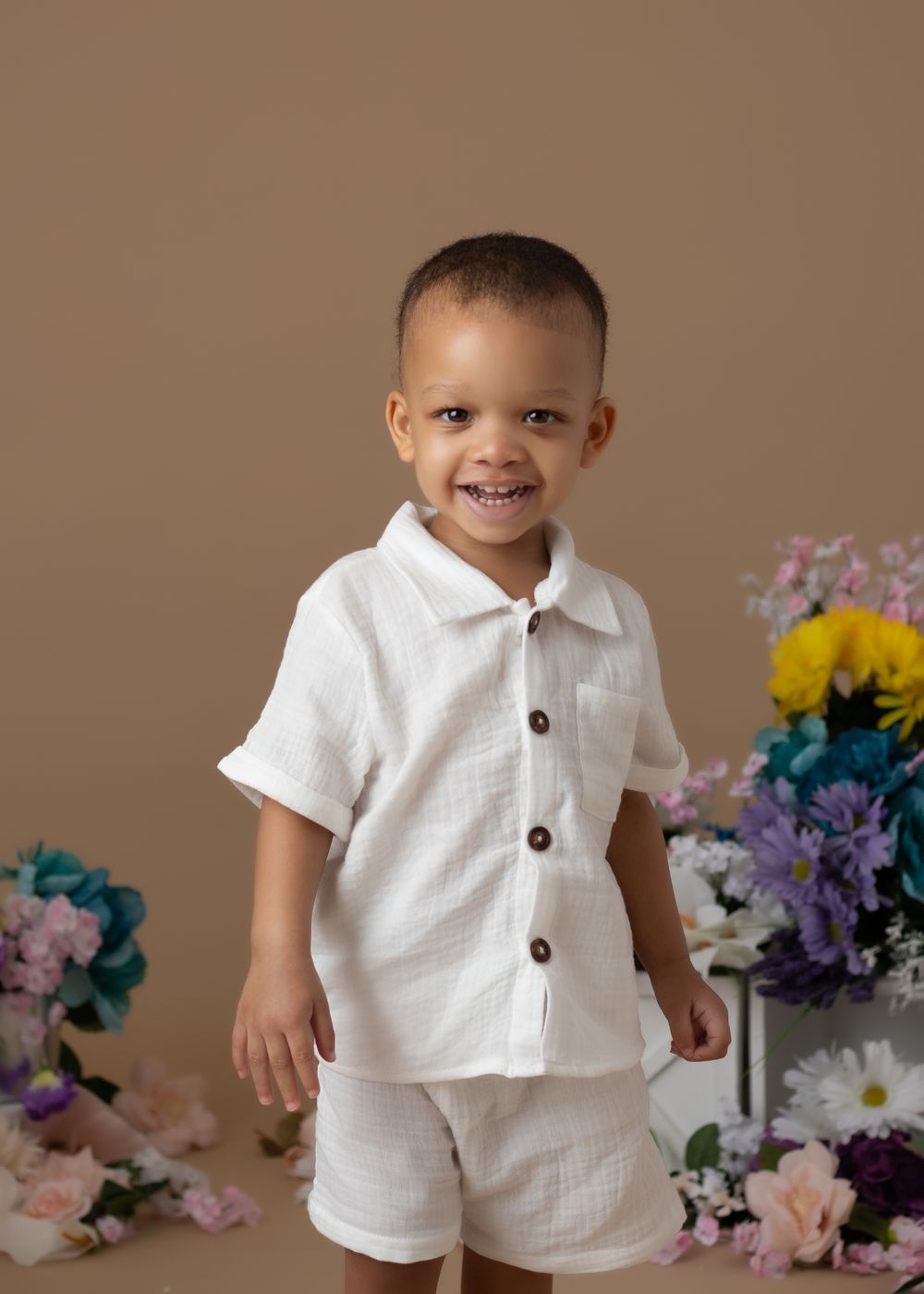 Smiling toddler boy in white linen shirt and shorts set, surrounded by colorful flowers on tan background.