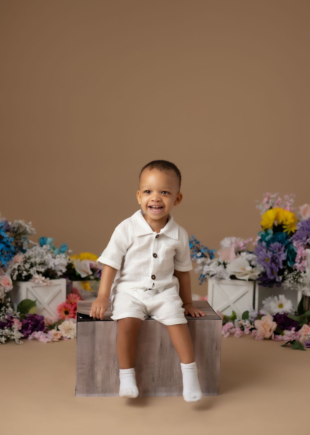 Smiling toddler boy in white outfit sitting on wooden box surrounded by colorful flowers on tan background.