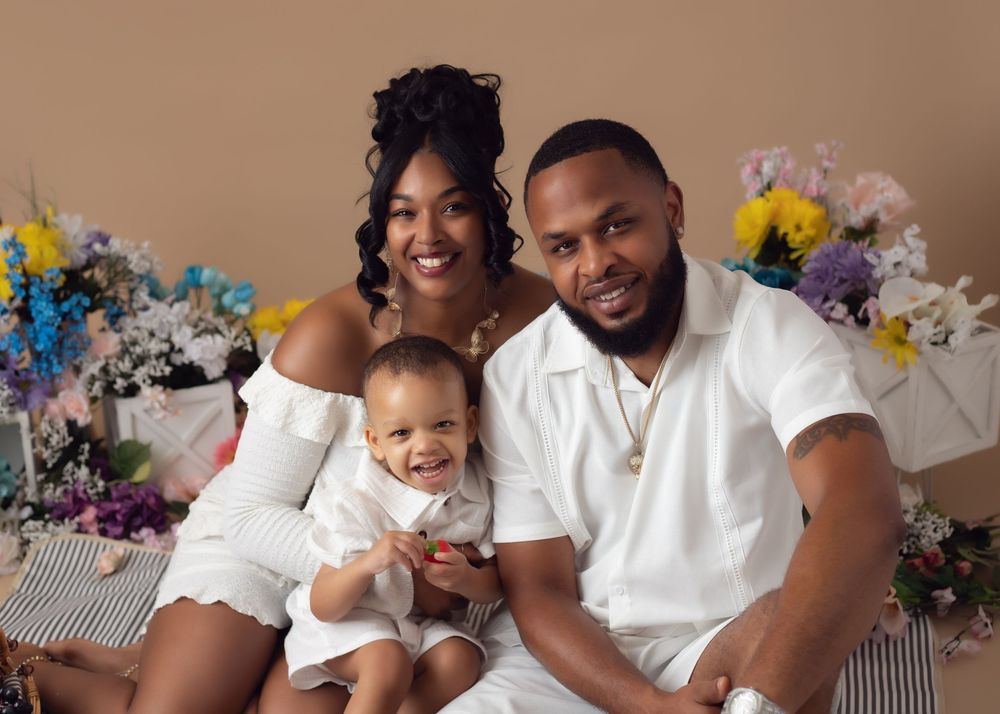 A happy family of three in white outfits poses with colorful flowers, parents smiling with their baby.