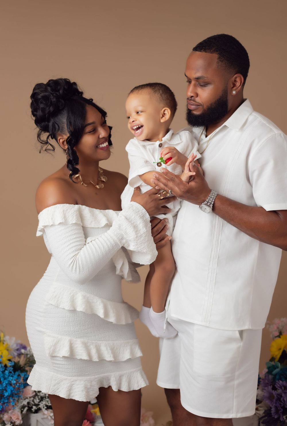 A joyful Black family in matching white outfits poses for a studio portrait, parents smiling at their laughing baby boy.