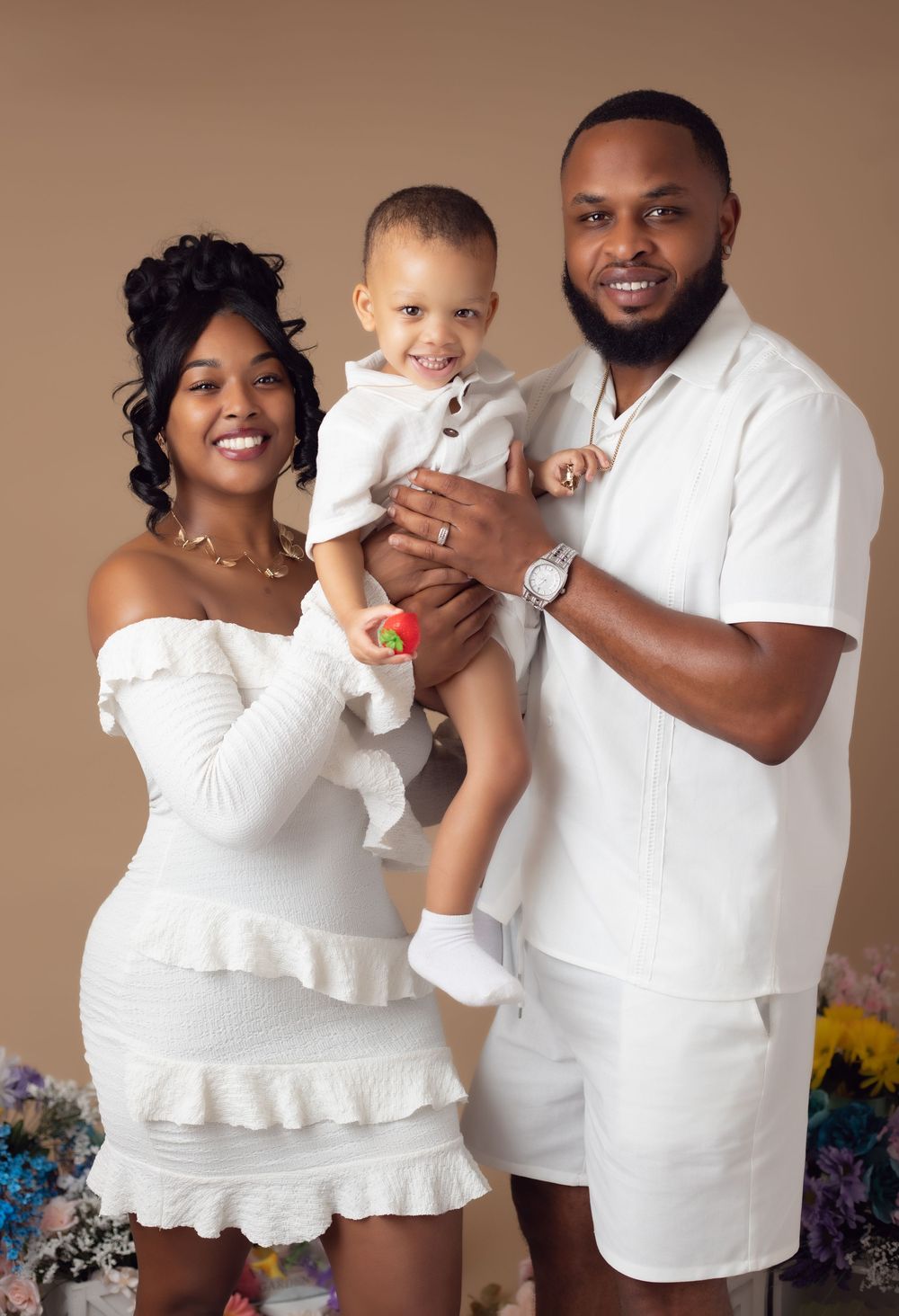 A joyful family of three in matching white outfits poses for a studio portrait against a warm beige background.