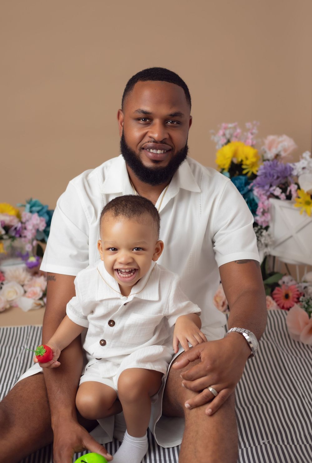 A man and toddler in matching white outfits smile together in a floral studio portrait.