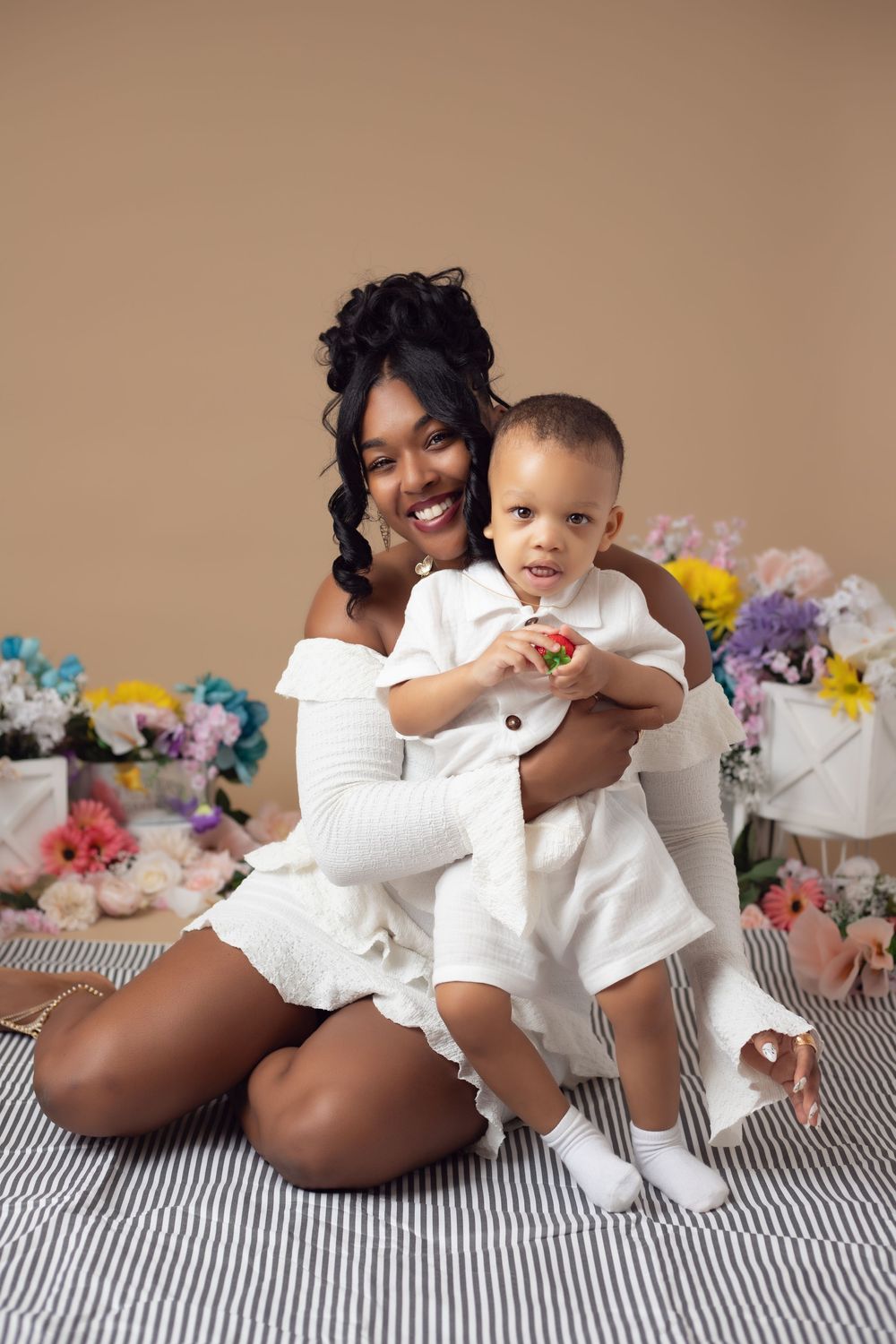 A smiling Black mother in white dress sits on striped floor holding her baby boy surrounded by colorful flowers.