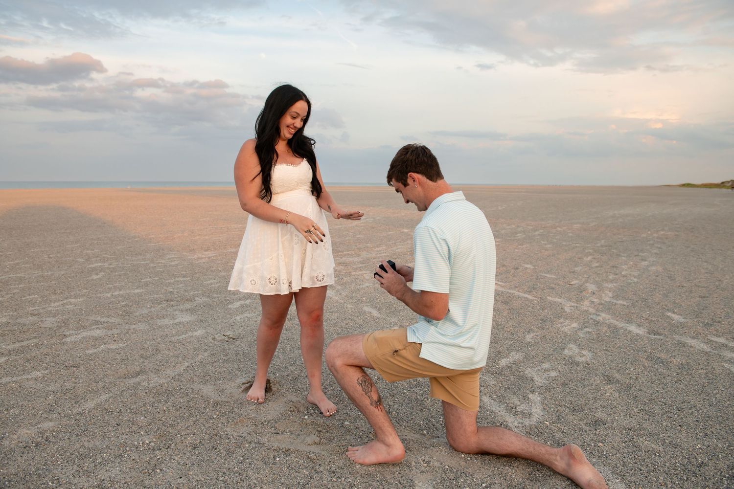 Man proposes on one knee to woman in white dress on sandy beach at sunset.