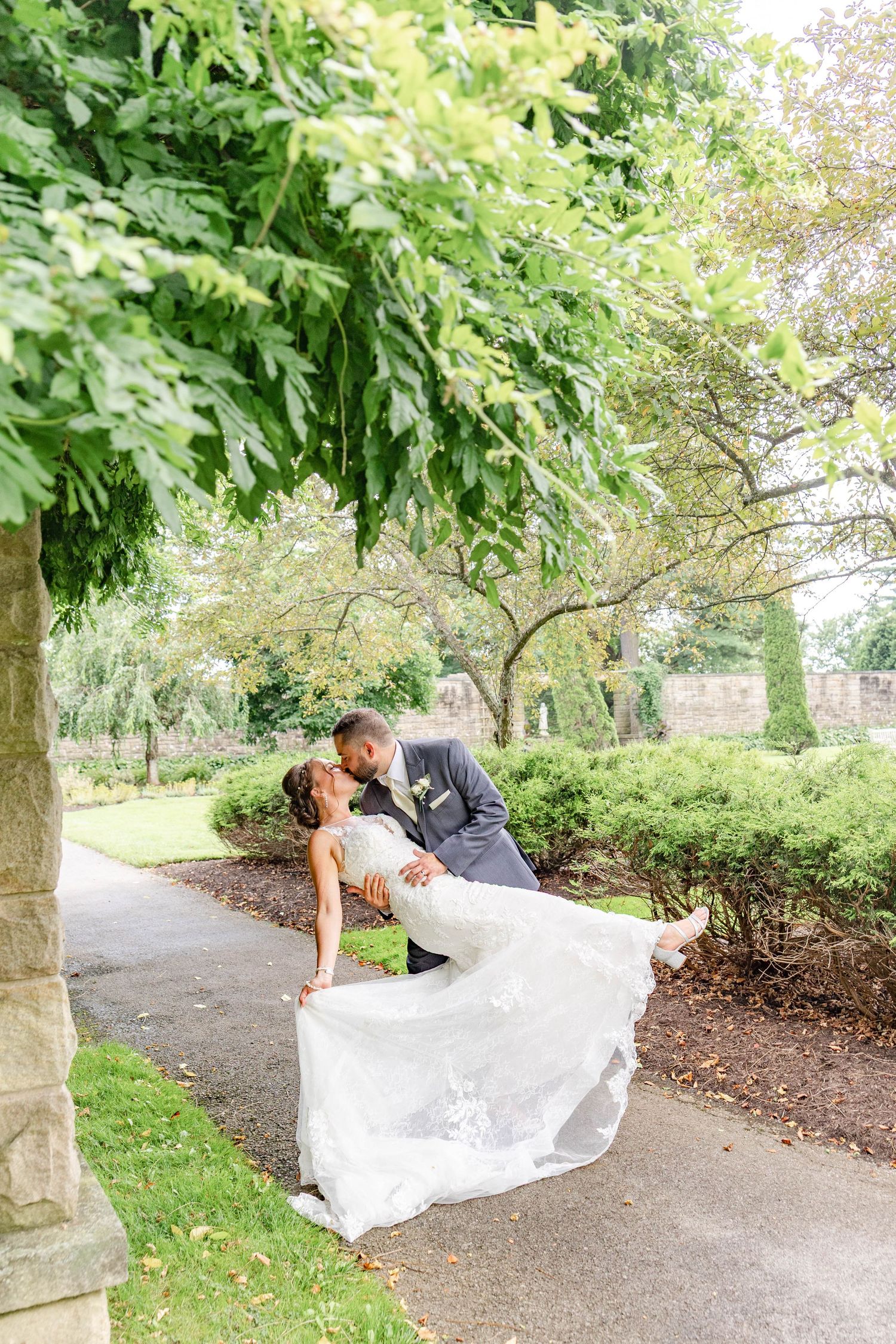Groom dips bride for a romantic kiss on a garden pathway surrounded by lush green trees on their wedding day.