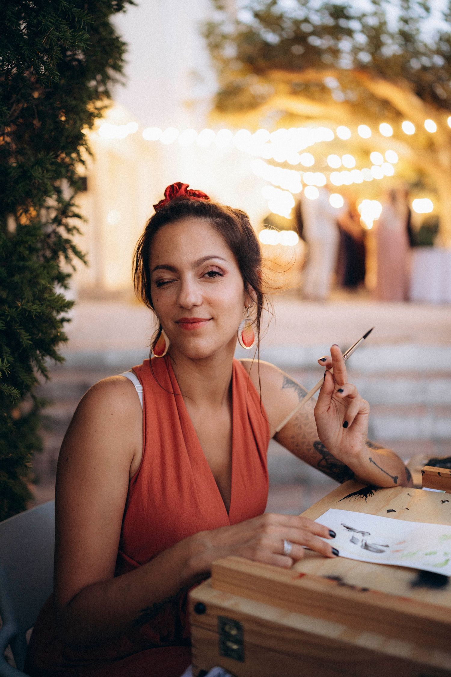 Smiling woman in orange dress sketching at outdoor cafe table with string lights in background.