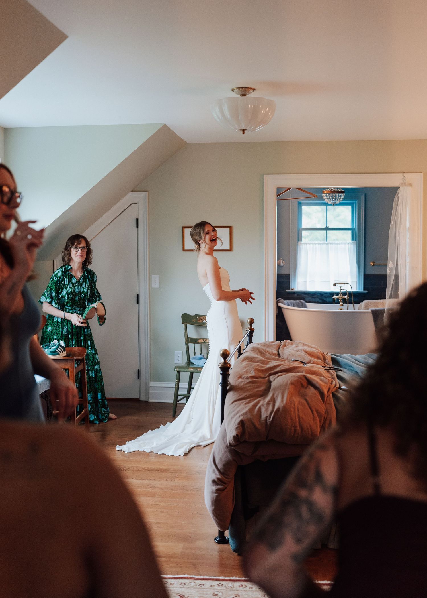 Bride in white wedding gown stands in rustic room as bridesmaids in green dresses look on admiringly.