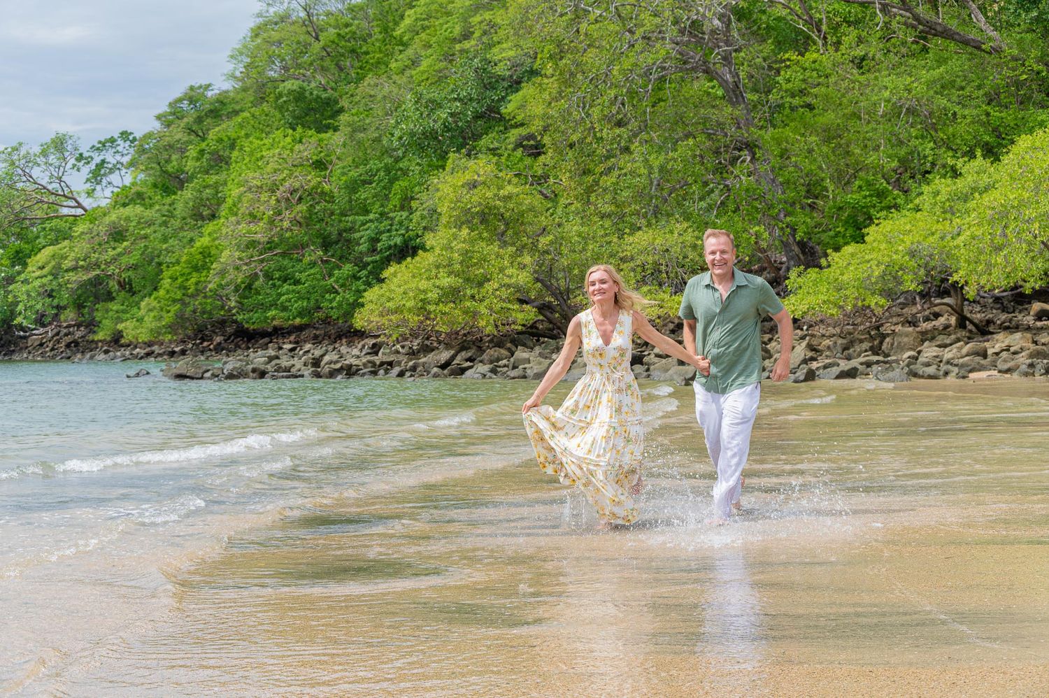 Couple holding hands running along a tropical beach with lush green trees and gentle ocean waves.