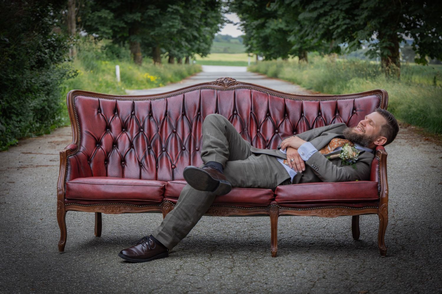 Groom in gray suit relaxes lying on a vintage red tufted sofa on a tree-lined road.