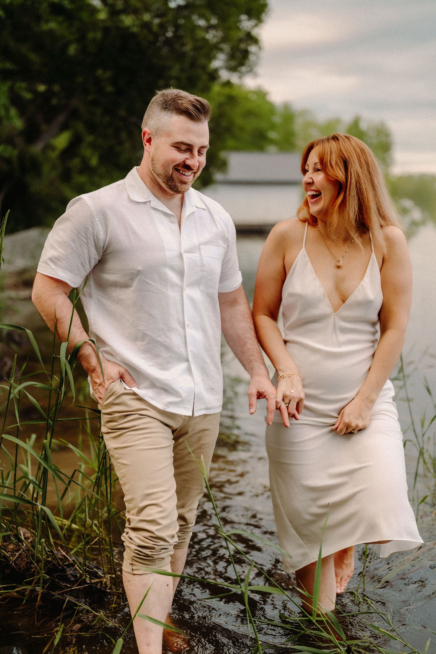 A smiling couple walks through shallow water, the man in a white shirt and the woman in a white slip dress, surrounded by greenery.