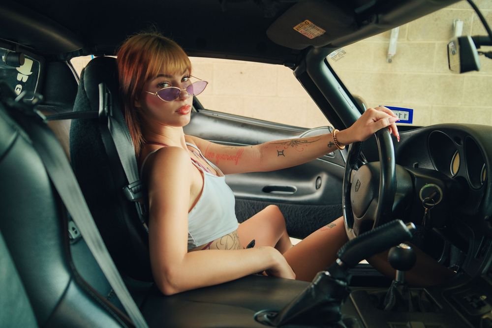 Young woman with pink hair and purple sunglasses sitting in car driver seat, hand on steering wheel.