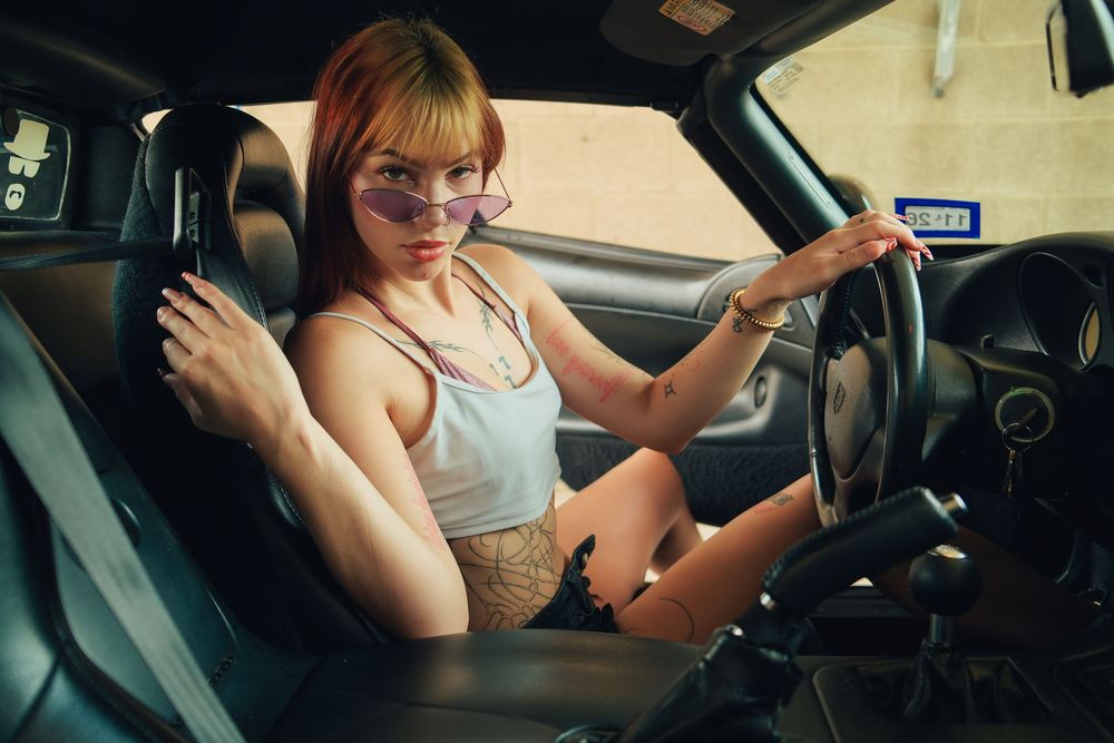 Young woman with tattoos and sunglasses sitting in car driver seat holding steering wheel, wearing gray tank top.