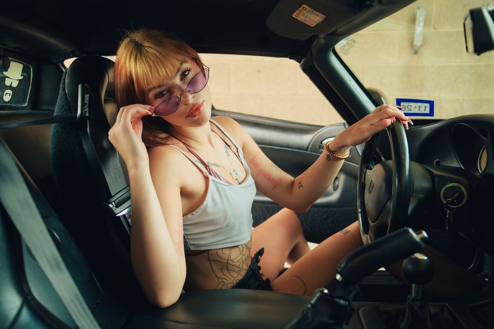 Young woman with tattoos and purple sunglasses sitting in car driver seat, wearing gray tank top.