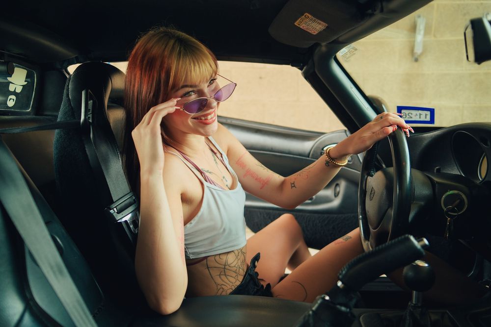 Young woman with tattoos and purple sunglasses sitting in car driver seat, wearing crop top and shorts.