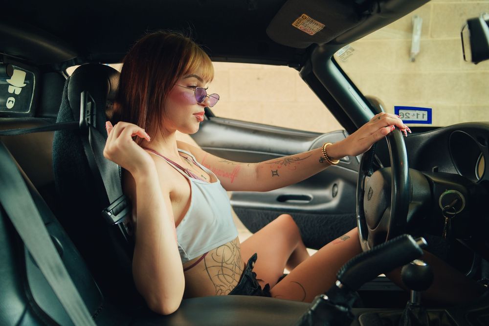 Young woman with tattoos and purple sunglasses sitting in car driver seat, wearing white top and denim shorts.