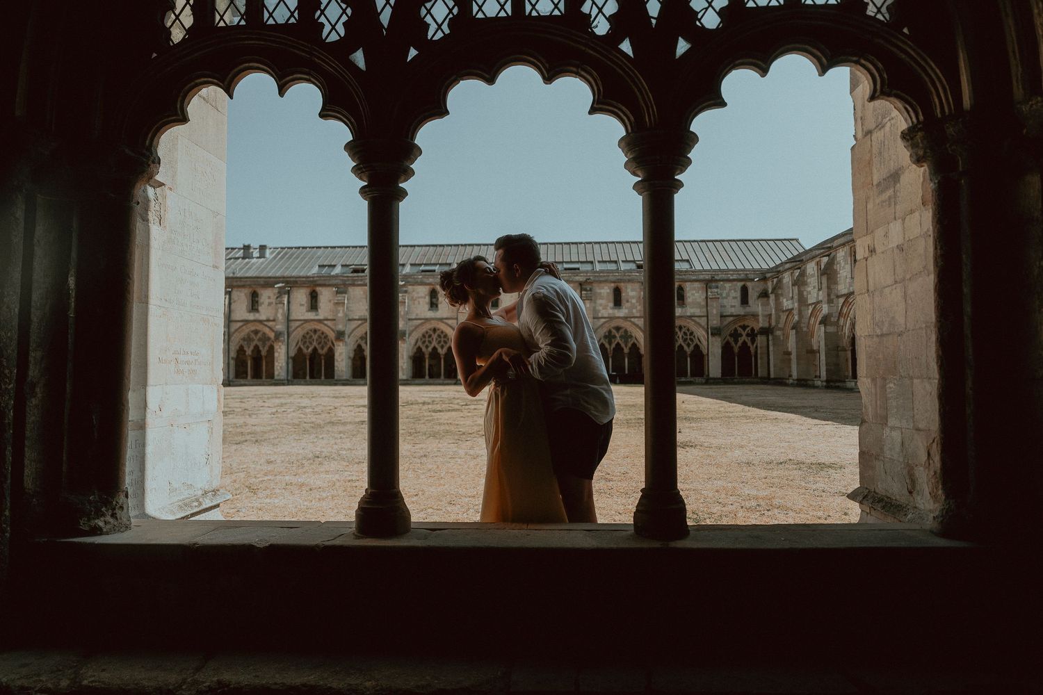 Couple sharing a romantic kiss framed by gothic arches in a sunlit historic courtyard.