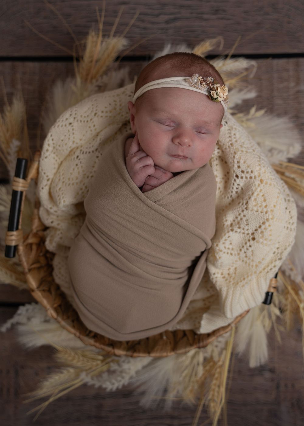 Sleeping newborn baby swaddled in tan wrap with gold headband in rustic wheat basket on wood floor.