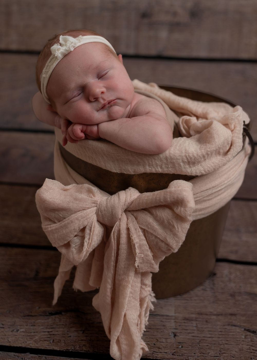 Sleeping newborn baby girl in a bucket wrapped in a soft peach muslin cloth with a bow.