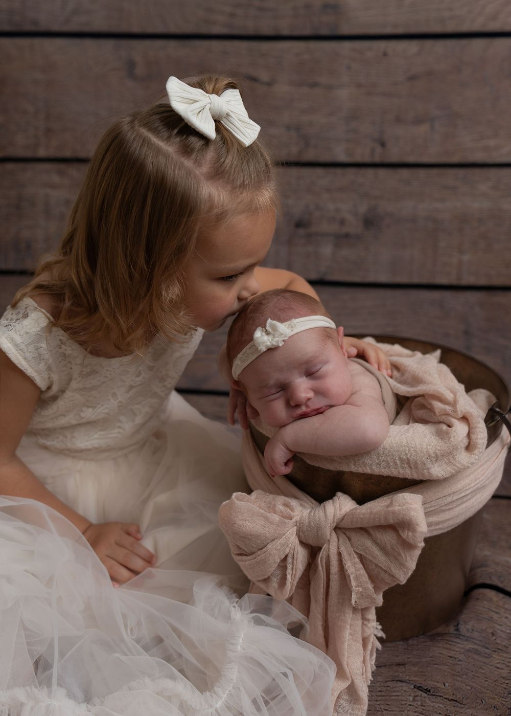 Toddler girl in white dress kissing her sleeping newborn sister in a bucket prop.