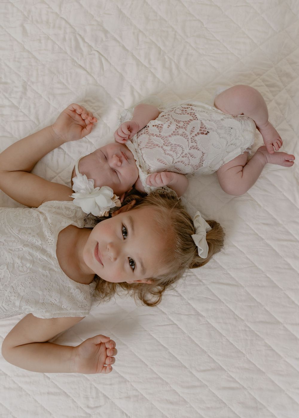 Overhead view of smiling toddler girl lying next to newborn baby sibling on white quilted blanket.