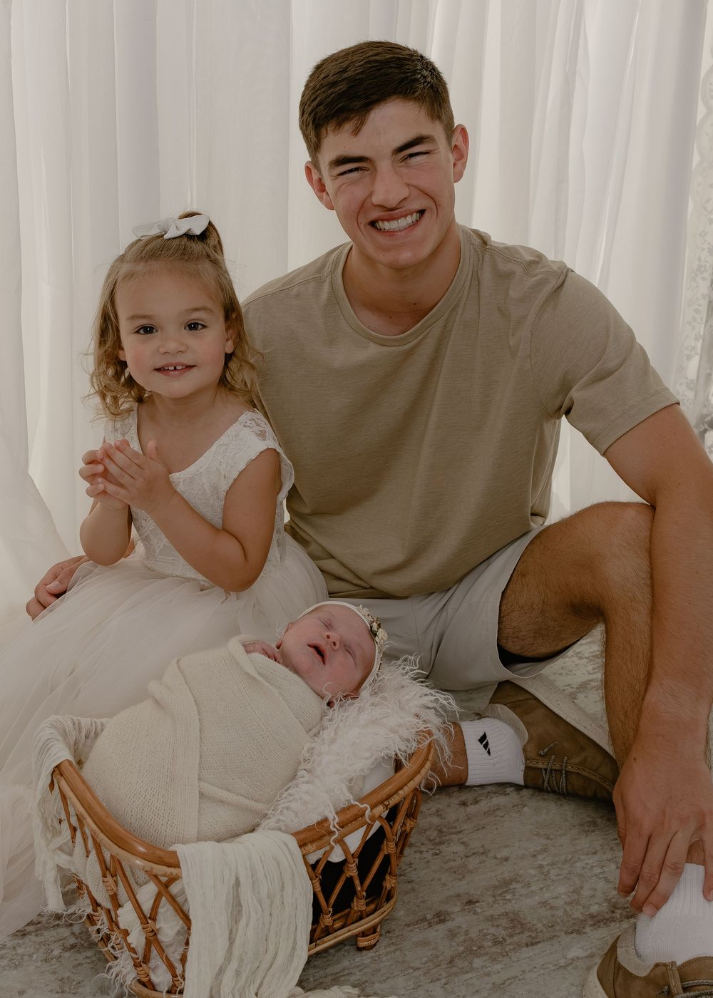 Smiling teenage boy poses with toddler girl and swaddled newborn baby in wicker basket indoors.