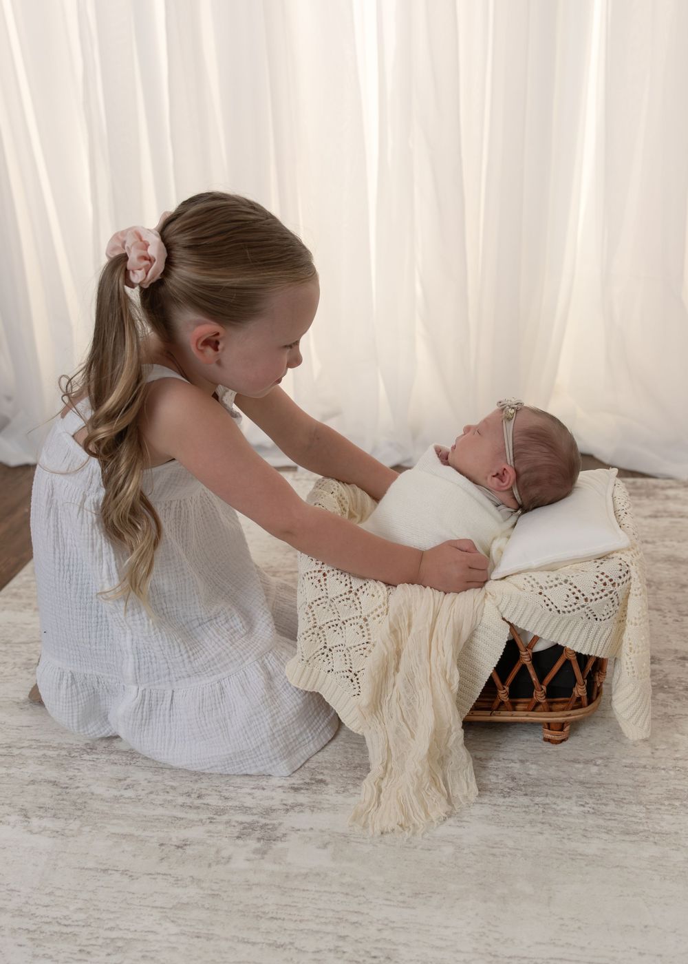 Young girl in white dress lovingly gazes at her newborn sibling in a wicker basket.